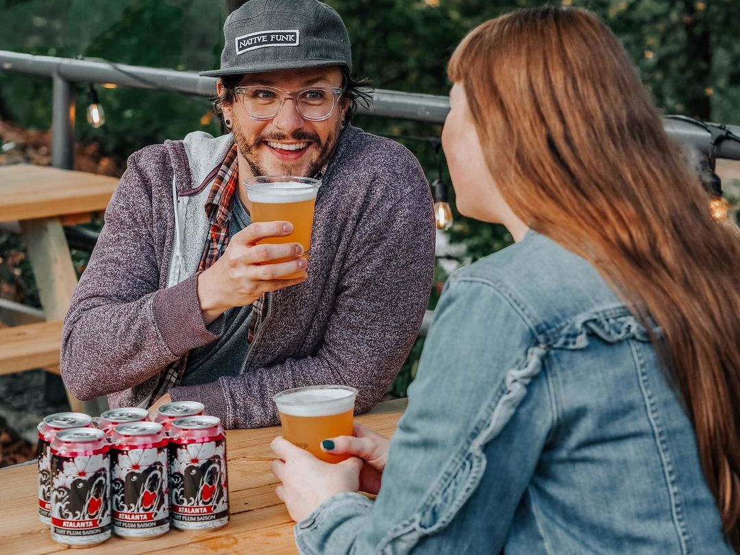 two people sitting across from each other and drinking a beer. Photo by Instagram user @orpheusbrewing