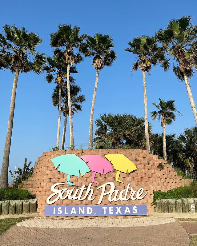 Palm trees and the South Padre Island sign with blue, pink, and yellow beach umbrellas. Photo by Instagram user @davidlanceres