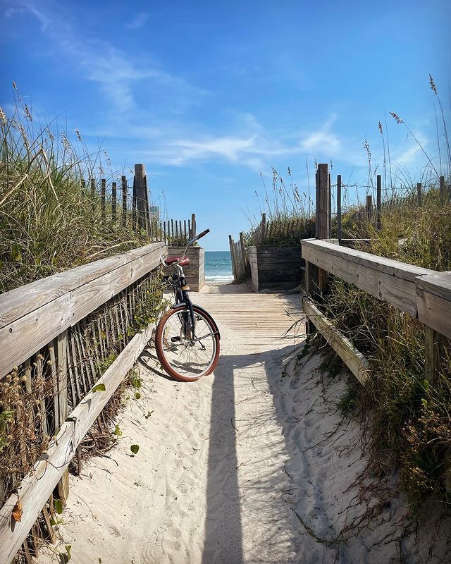 Sandy beach boardwalk surrounded by tall green grass. Photo by Instagram @ka_bliss