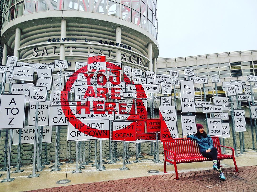 A women sits on a street bench with an art display of signs behind her reading you are here. Photo by Instagram user @trek_n_stef