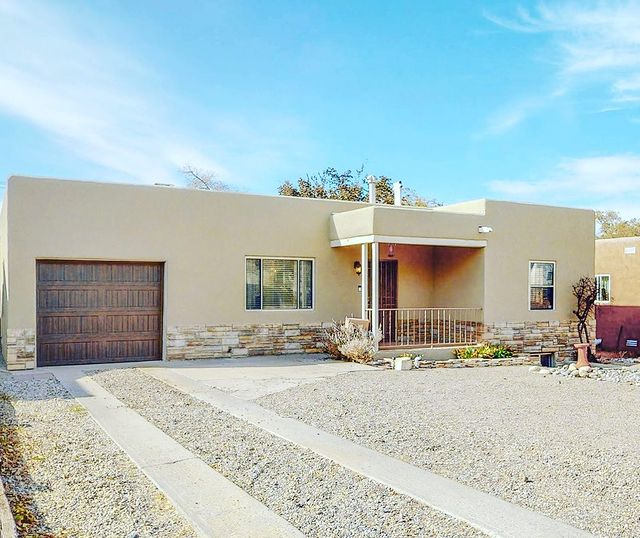 A one-story adobe home with a wooden door and gravel yard, plus a few small bushes and shrubs near the front covered porch on a partly cloudy day. Photo via Instagram user @albuquerquehomes