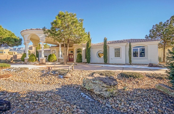 An elaborate spanish revival style home with a gravel front yard, tall trees and shrubs, and mountains rising in the distance beneath a clear blue sky. Photo via Instagram use @newmexicohomegroup