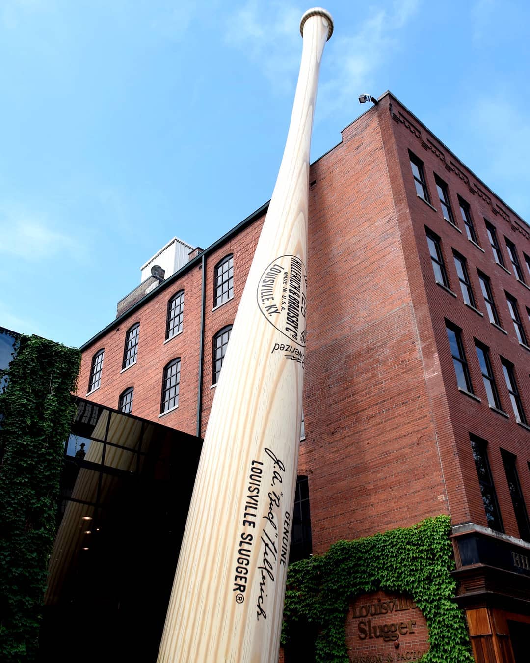 Looking up at a giant bat statue that leans against big red brick Louisville Slugger Museum & Factory. Photo by Instagram user @styleblueprint