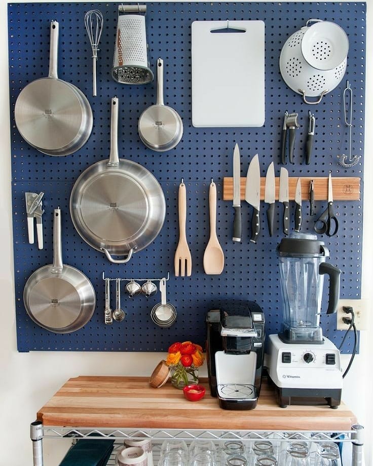 a blue pegboard hanging on the wall with kitchen utensils, pans, a magnetic knife holder, and a cutting board hanging on it