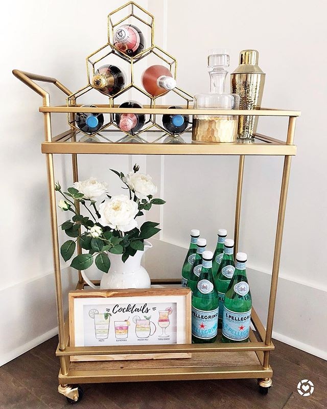 a gold bar cart sitting in the corner with wine bottles and a cocktail shaker on the top rack and a vase with flowers and more bottles on the bottom rack