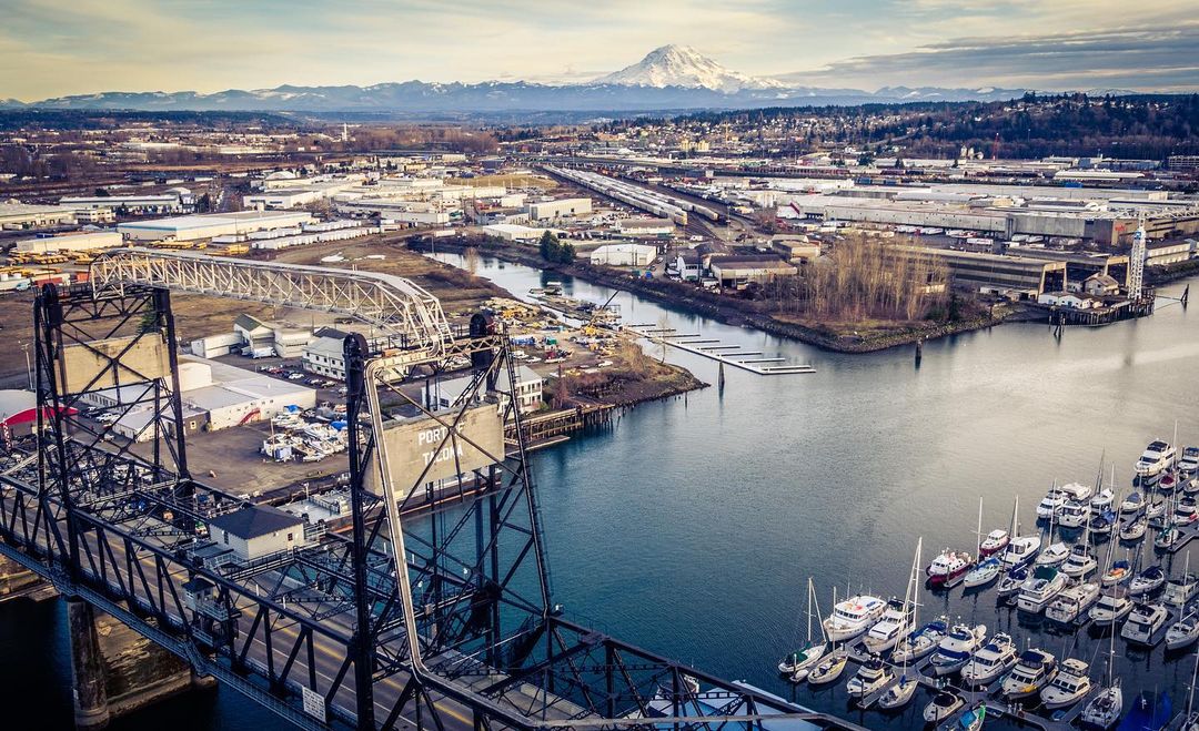 Drone photo of Tacoma Washington with industrial bridge, sailboats, and water in foreground with snowy mountain and town in background | Photo by Instagram user @graymediapro