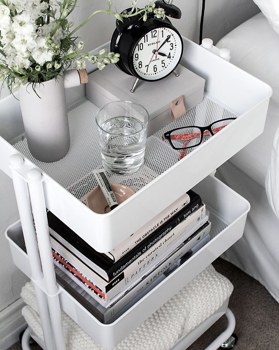 White rolling storage cart filled with decor next to bed. Photo by Instagram user @housejunkie_