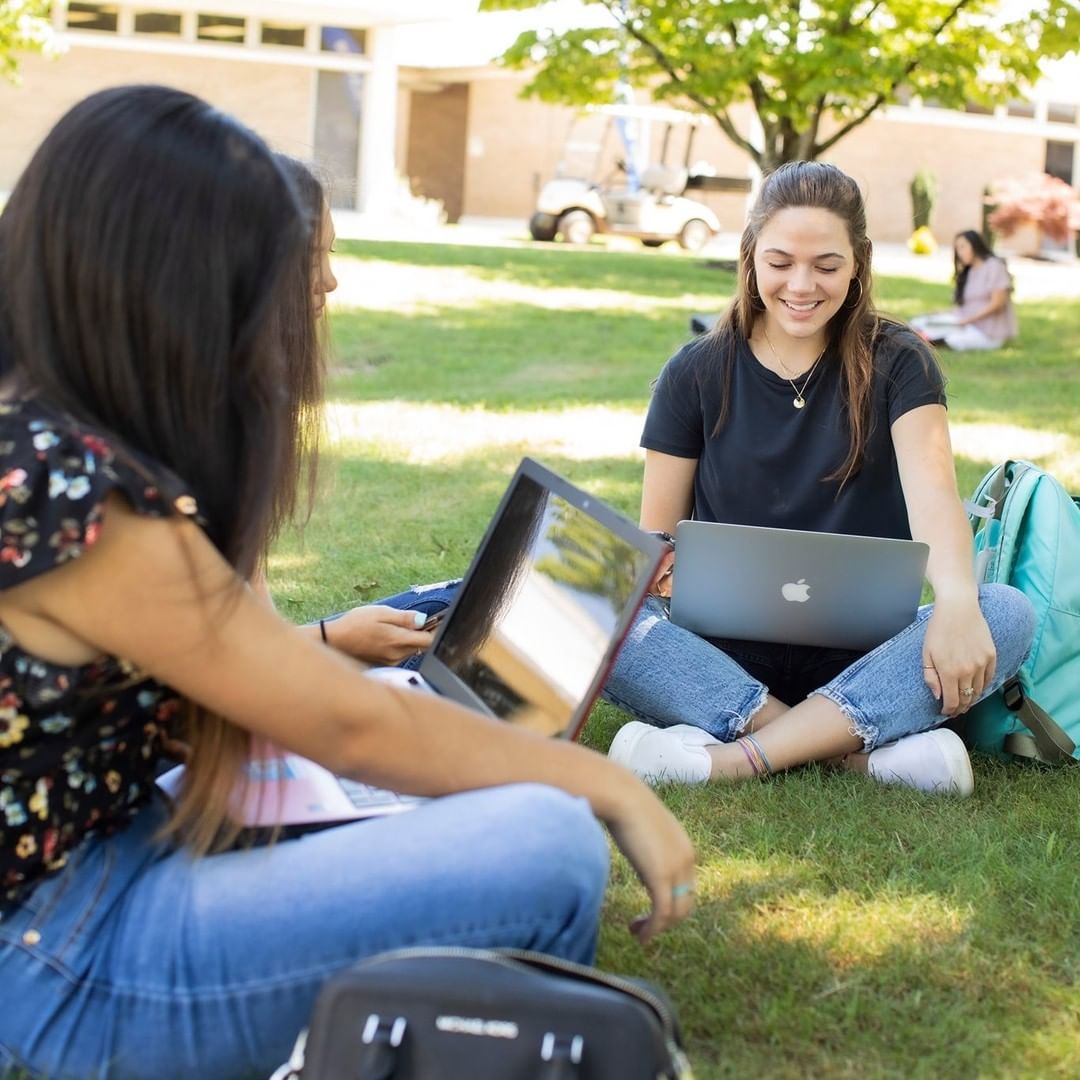 Two students sitting on the grass outside working on laptops. Photo by Instagram user @northeastalcommunitycollege
