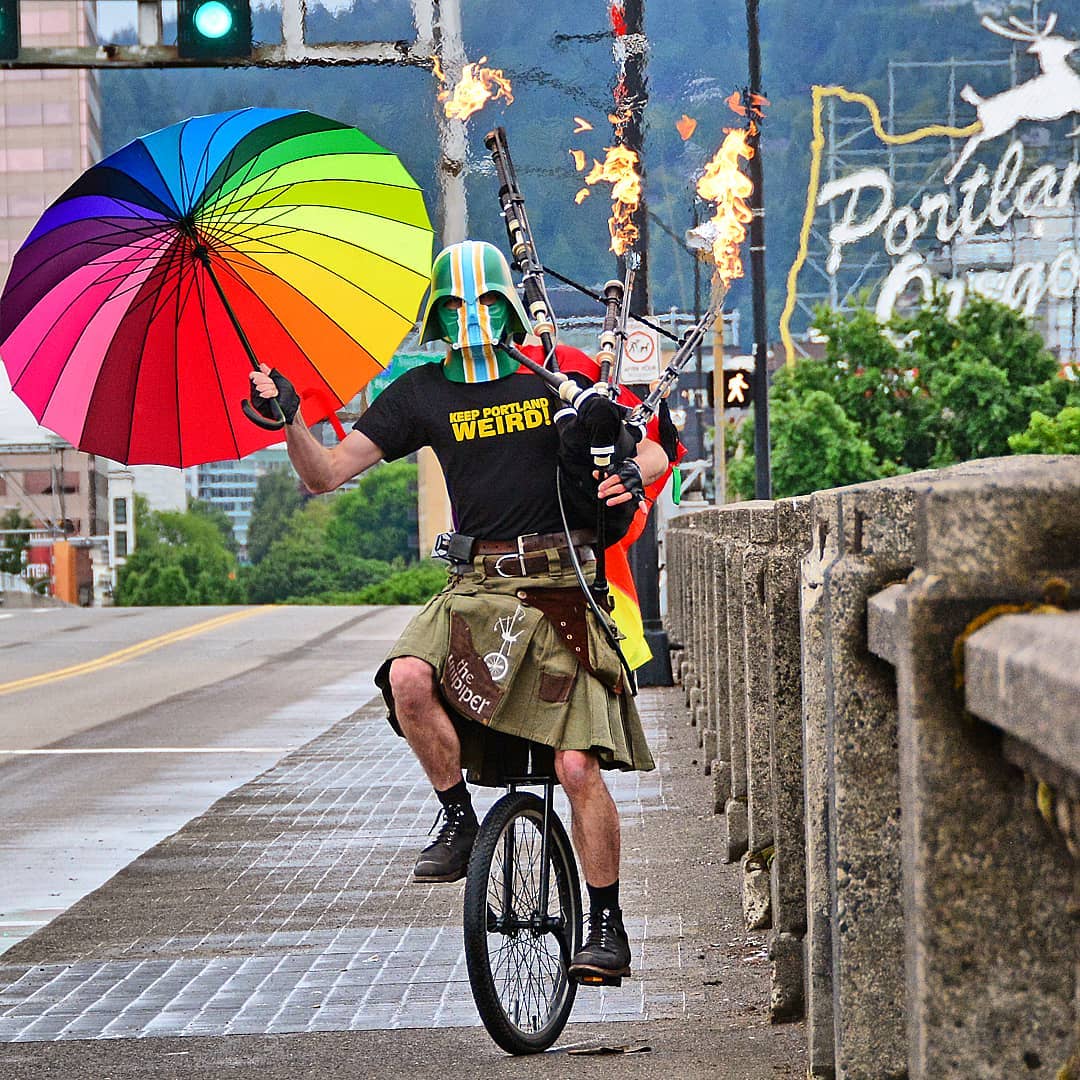 Man on a unicycle wearing a kilt, holding a colorful umbrella and a flaming bagpipe wearing a colorful Darth Vadar mask Photo by Instagram @theunipiper
