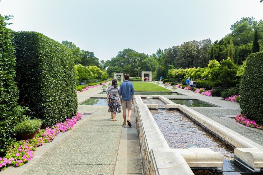 Two people stroll hand in hand on a sidewalk between a concrete pool with water on the right and shrubs on the left in a sunny garden. Photo via Instagram user @visit_dallas