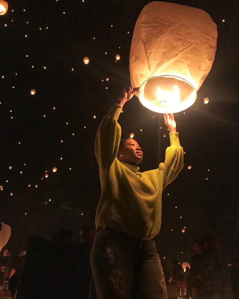 A woman holds a lit lantern while the dark sky behind her is lit with flying lanterns scattered like stars. Photo via Instagram user @so.phone.knee