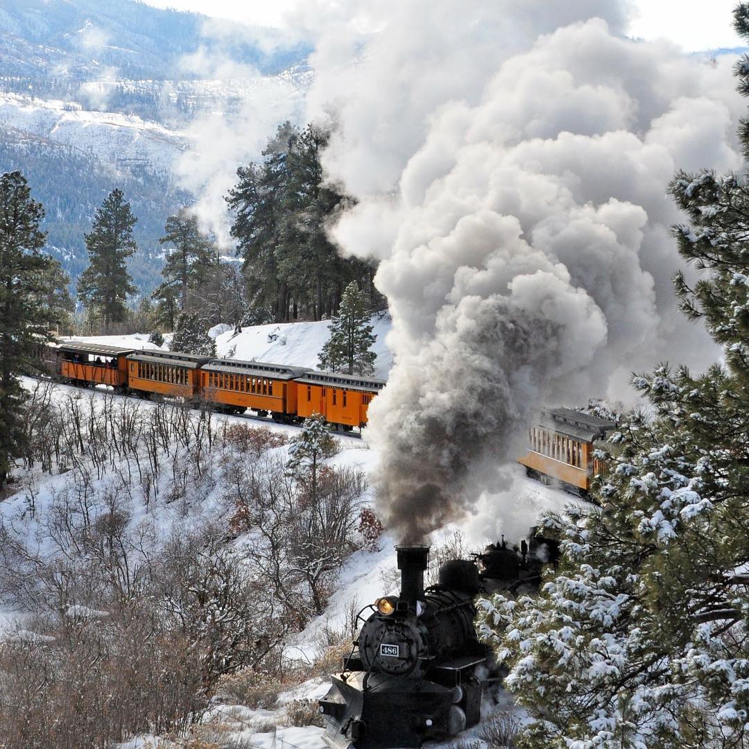Orange train riding through snowy mountains. Photo by Instagram user @lindyloumomof2 