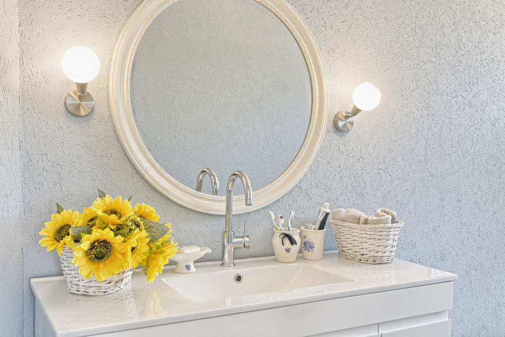 A white mirror above a sink with white countertops and a basket of sunflowers
