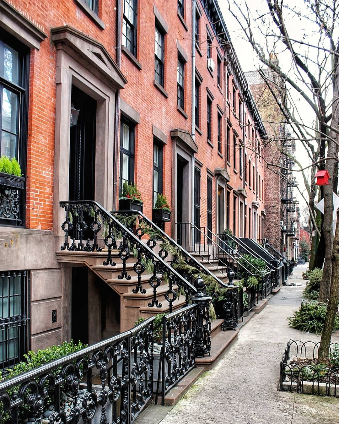 Sidewalk view of multiple red brick brownstones with black detailed railings. Photo by Instagram user @jonathankamensky