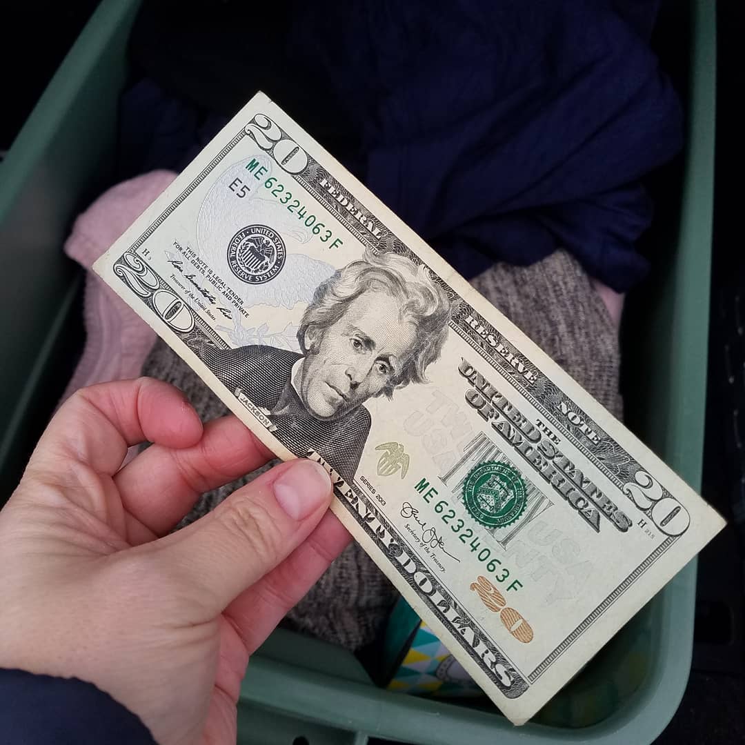 Hand Holding $20 Bill with Hamper of Old Clothes in Background. Photo by Instagram user @ksbudgetmom