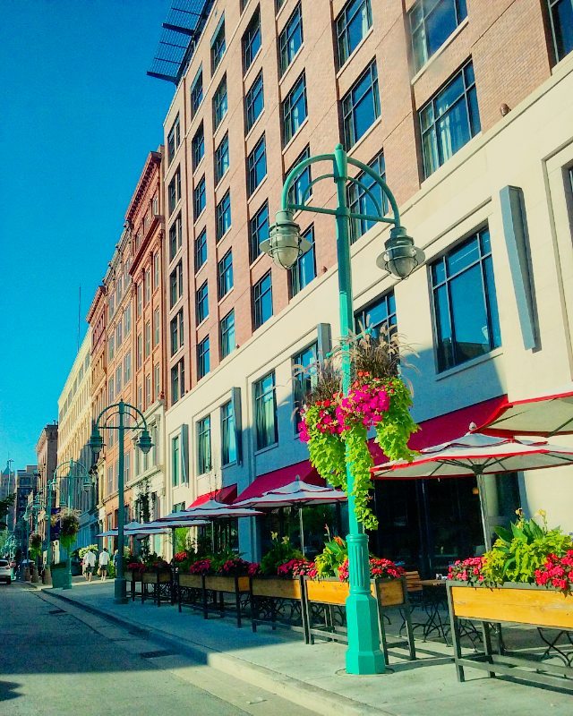 Tall building sits on a city street as street lights and patio tables line the sidewalk from businesses. Photo by Instagram user @debskaleidoscope