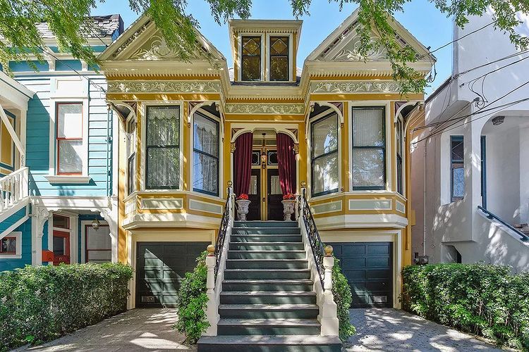 Exterior shot of yellow Victorian house with white trim and black garage doors and walk-up stairs that lead to front doors with red curtain in Noe Valley San Francisco. Photo by Instagram user @noevalleysf