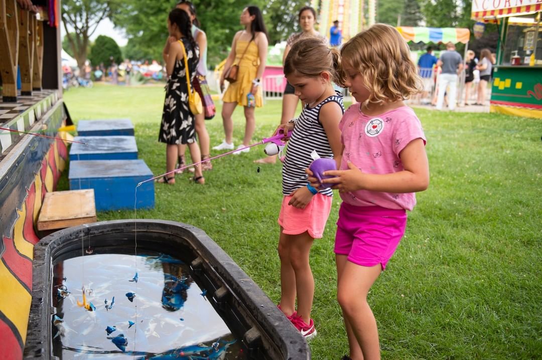 Photo of two girls wearing pink shorts and striped shirts fishing for plastic sharks in water trout during carnival days for Monona Festival in Madison, WI. Photo by Instagram user @mononafestival