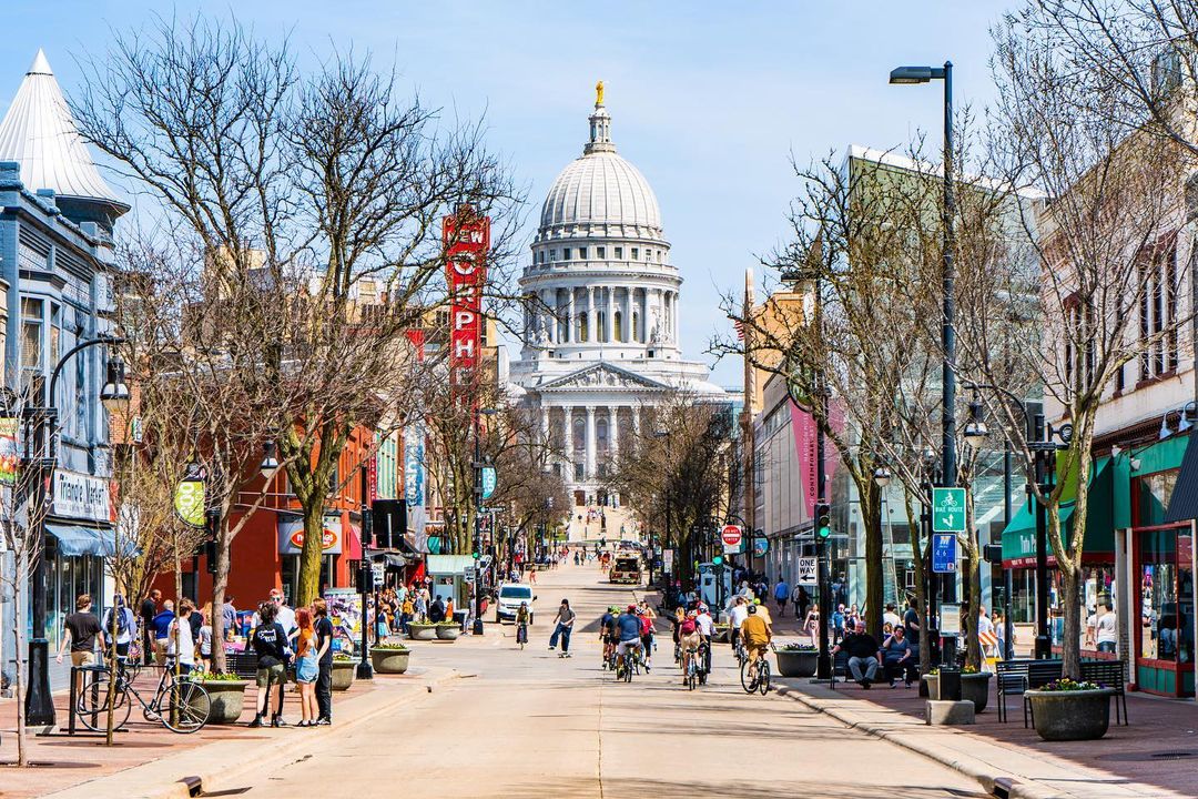Photo on State Street in Madison looking east at Wisconsin State Capitol in the background with cyclists on street and people on sidewalks around local businesses. Photo by Instagram user @theonlyvimal