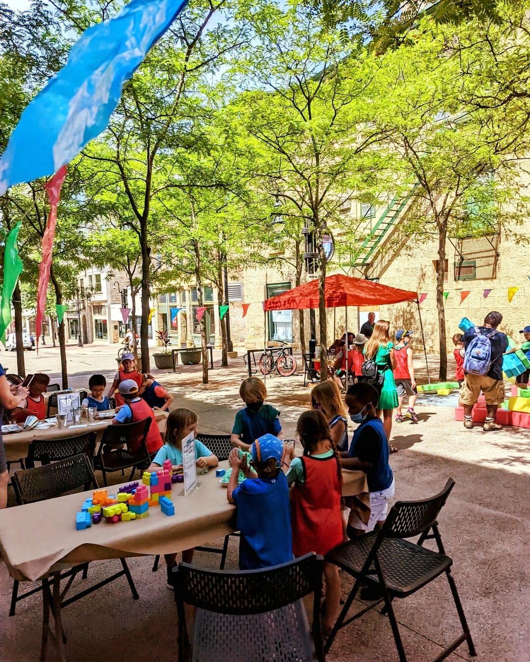 Photo of children playing with building blocks and making crafts on tables set up on State Street in Downtown Madison, WI in shade during pop-up family event. Photo by Instagram user @visitdowntownmadison 
