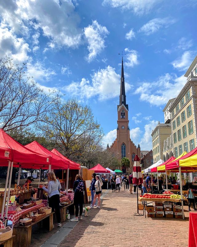 A brick paved road, blue skies with white fluffy clouds, and trees with new green buds. A church with a tall steeple in the background, and a farmer's market along the sides of the brick road featuring plenty of fresh fruits and vegetables as well as happy customers. Photo via Instagram user @walkablecharleston