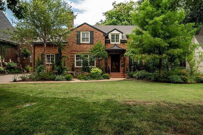Two story home with dark red brick and dark colored shutters, trim and roof lined with greenery and a tall tree in the front yard. Photo by Instagram user @theourengroup
