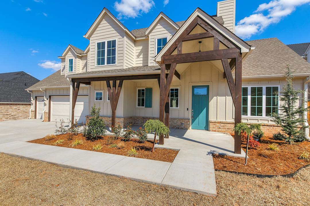 Two story modern farmhouse with beige siding and brick and tall wooden front porch over hang and a teal front door. Photo by Instagram user @michellemassiehomes