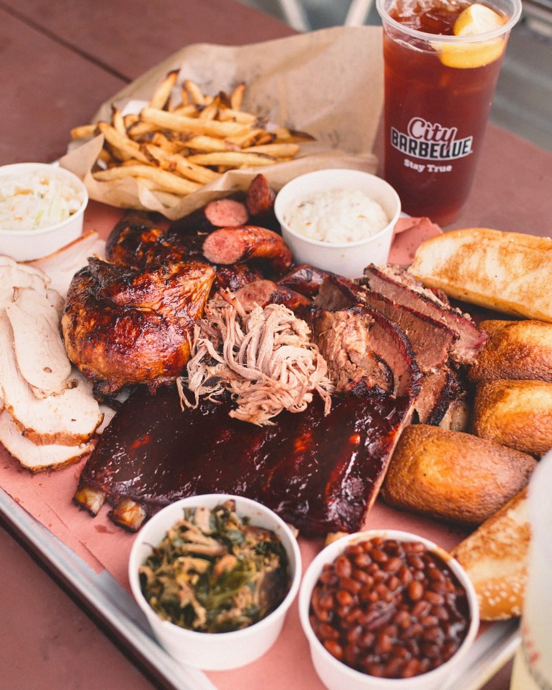 Overhead view of large tray of barbeque meats, cornbread, sides, and a glass of iced tea. Photo by Instagram user @citybbq