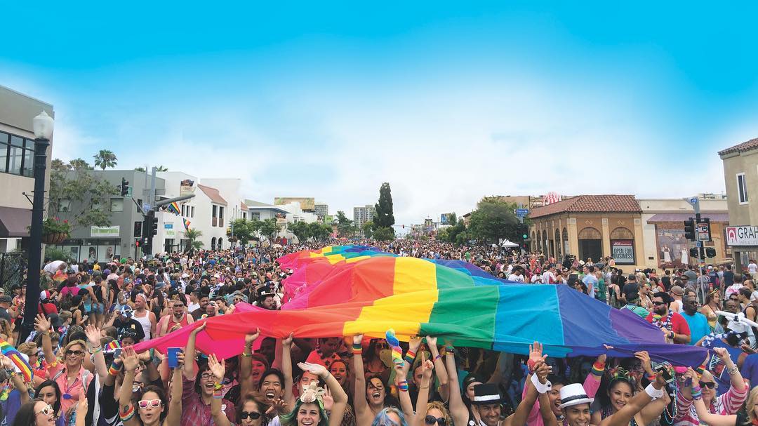 A large group of people stand on the street holding a giant rainbow flag above their head. Photo by Instagram user @sandiegopride