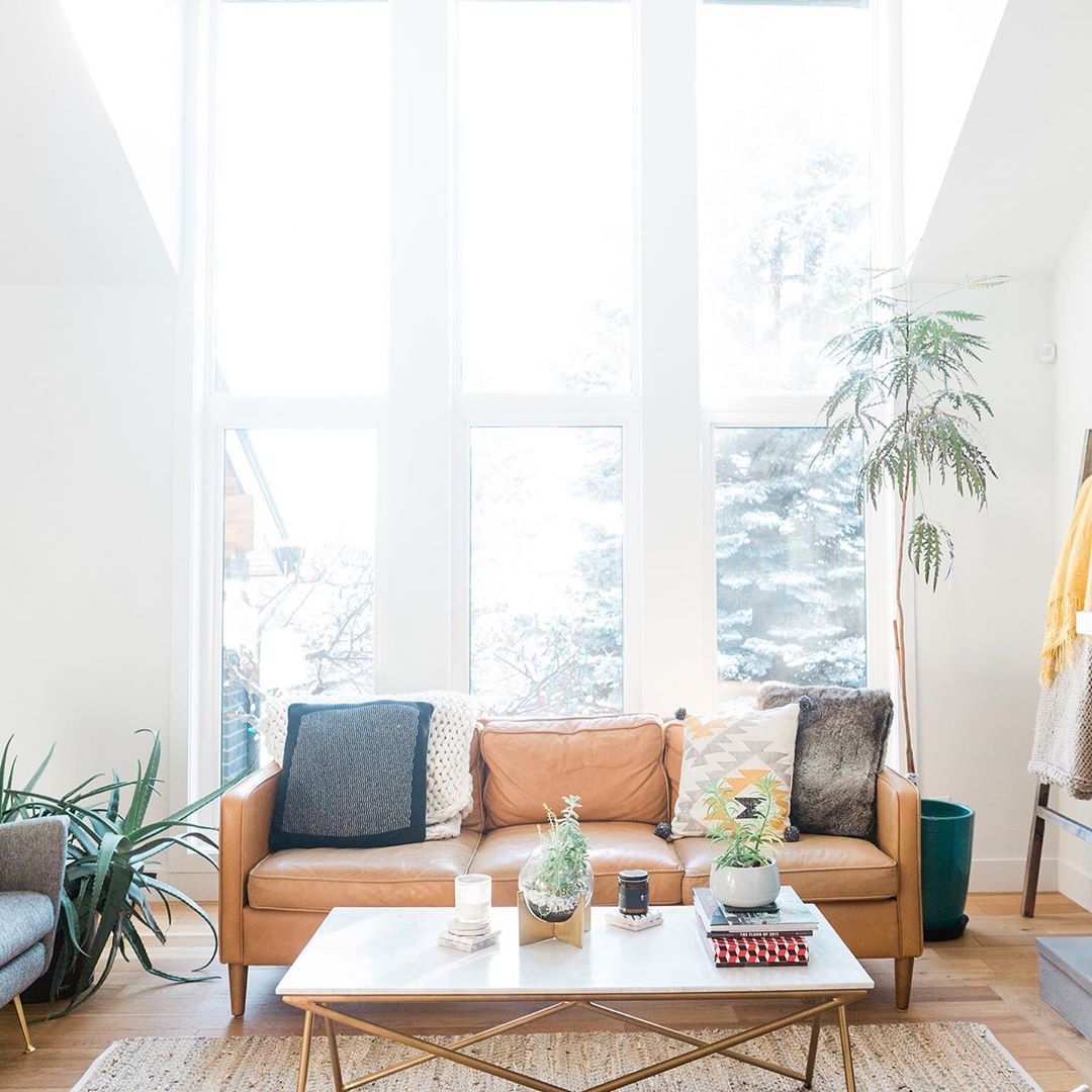 Living room with big windows and light brown leather couch with patterned pillows. Photo by Instagram user @move.play.mom