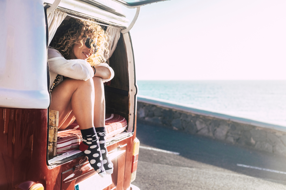Girl sitting out of the back of a van near the ocean.