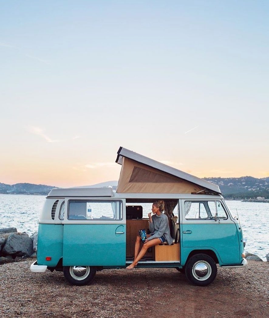 Girl sitting in the door of her blue van by the ocean. Photo by Instagram user @excampingex