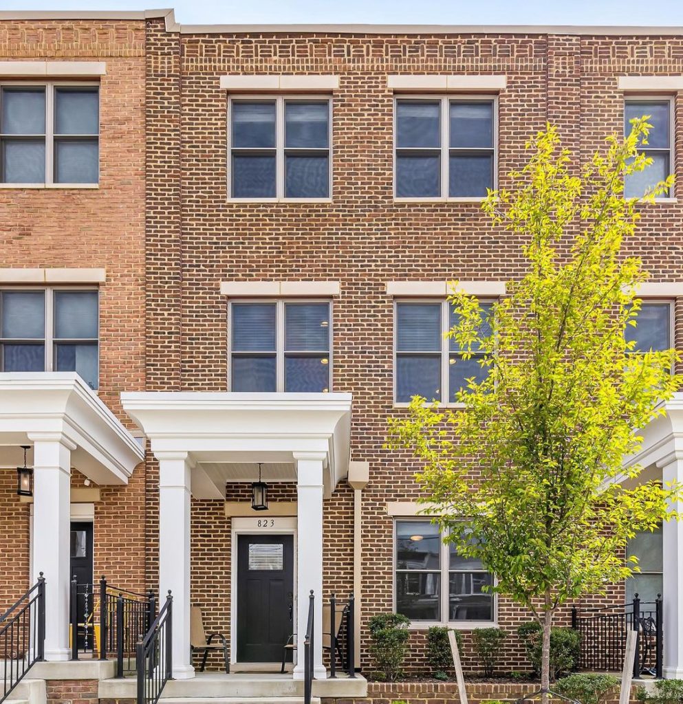 Plain, square, brick rowhouses with 4 paned windows and white pillared columns holding up an overhang over the front door. Small tall tree on right side. Photo via Instagram user @carolinekuntz_homes