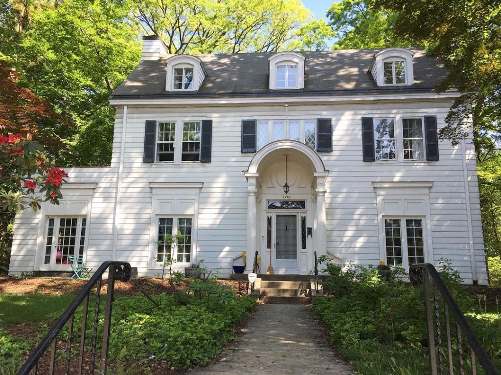 Large white colonial home with arch over doorway, black window shutters and roof. Lush, but short bushes line the sidewalk with stairs leading up from the street to the house. Large trees shadow the house and fill the sky in the background. Photo via Instagram user @joangoldmanrealtor