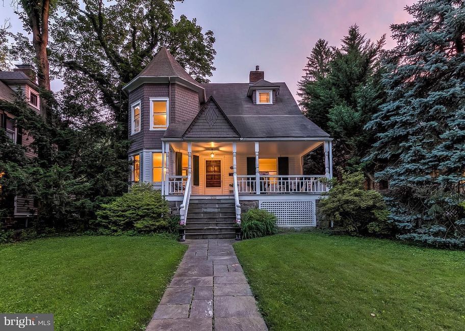 Small Victorian Queen Anne style home with a touret on left, small upstairs window on right roof, and white paint details making the brown wood features stand out. It's twilight and the porch lights light up the home amidst evergreen trees, bushes, and a tidy lawn. Photo via Instagram user @baltfishbowl