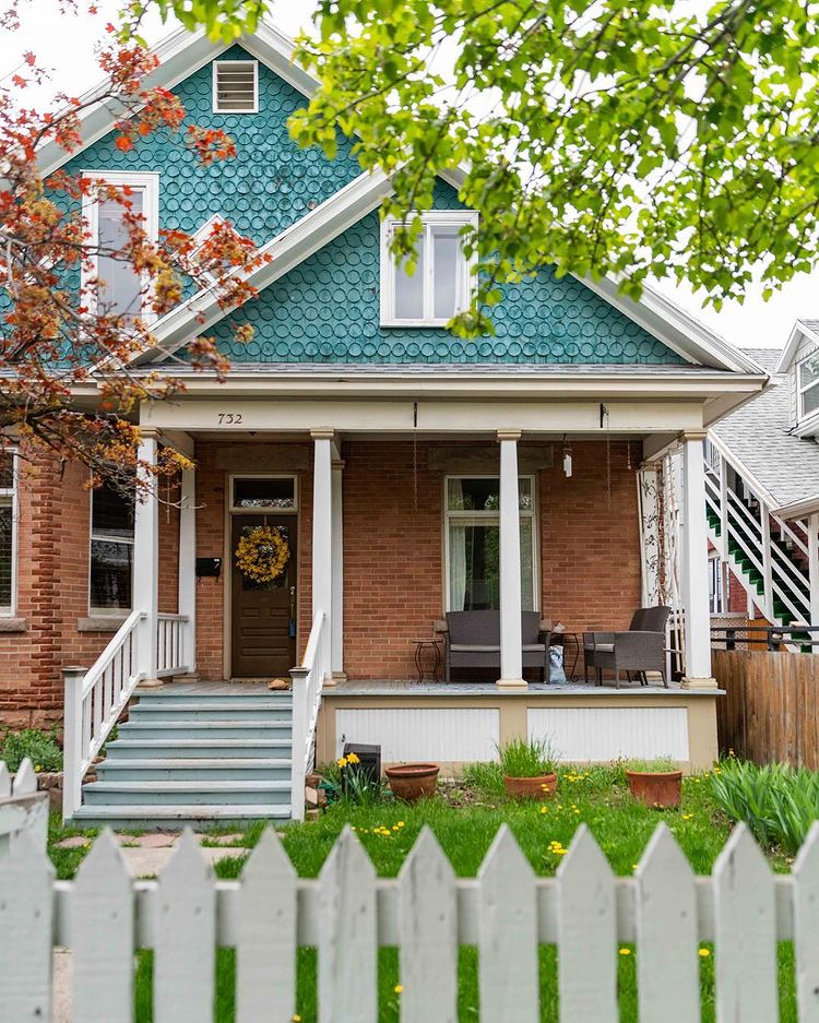 A brick bungalow with a blue walled attic, white pillars and picket fence, and a green lawn. Photo via Instagram user @thevictorianbungalow