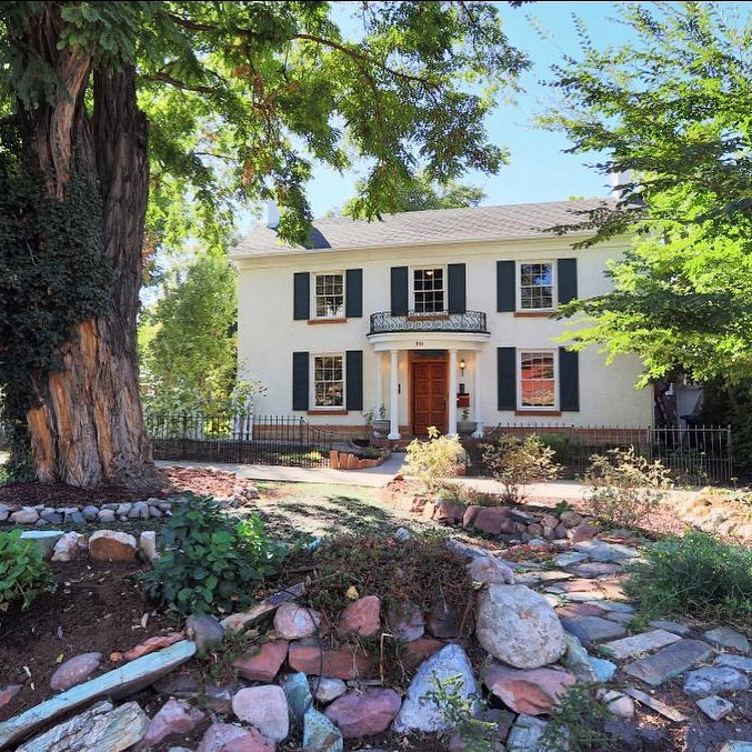 A white colonial home with black accents sits in the background of a rocky landscaped yard with trees, bushes, and brick paths. Photo via Instagram user @realtormarshall