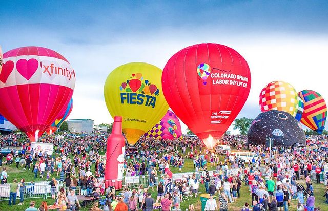 Several hot-air baloons of varying colors are being sent in to the air for the Labor Day Liftoff event while a huge crowd of people watch. @labordayliftoff