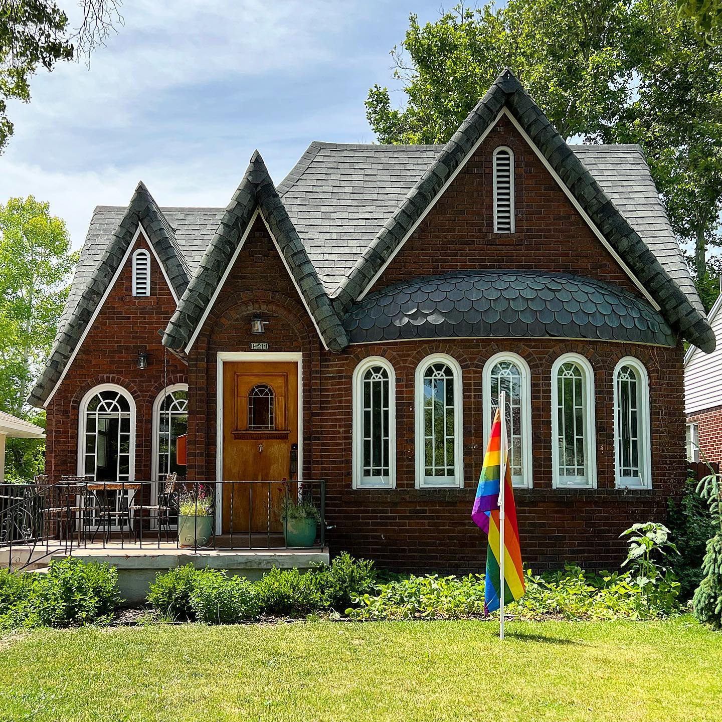 A dark red brick cottage with black shingles, white accents on windows and doors, and a LGBT pride flag in the front yard. Photo via Instagram user @lovelyoldhomes
