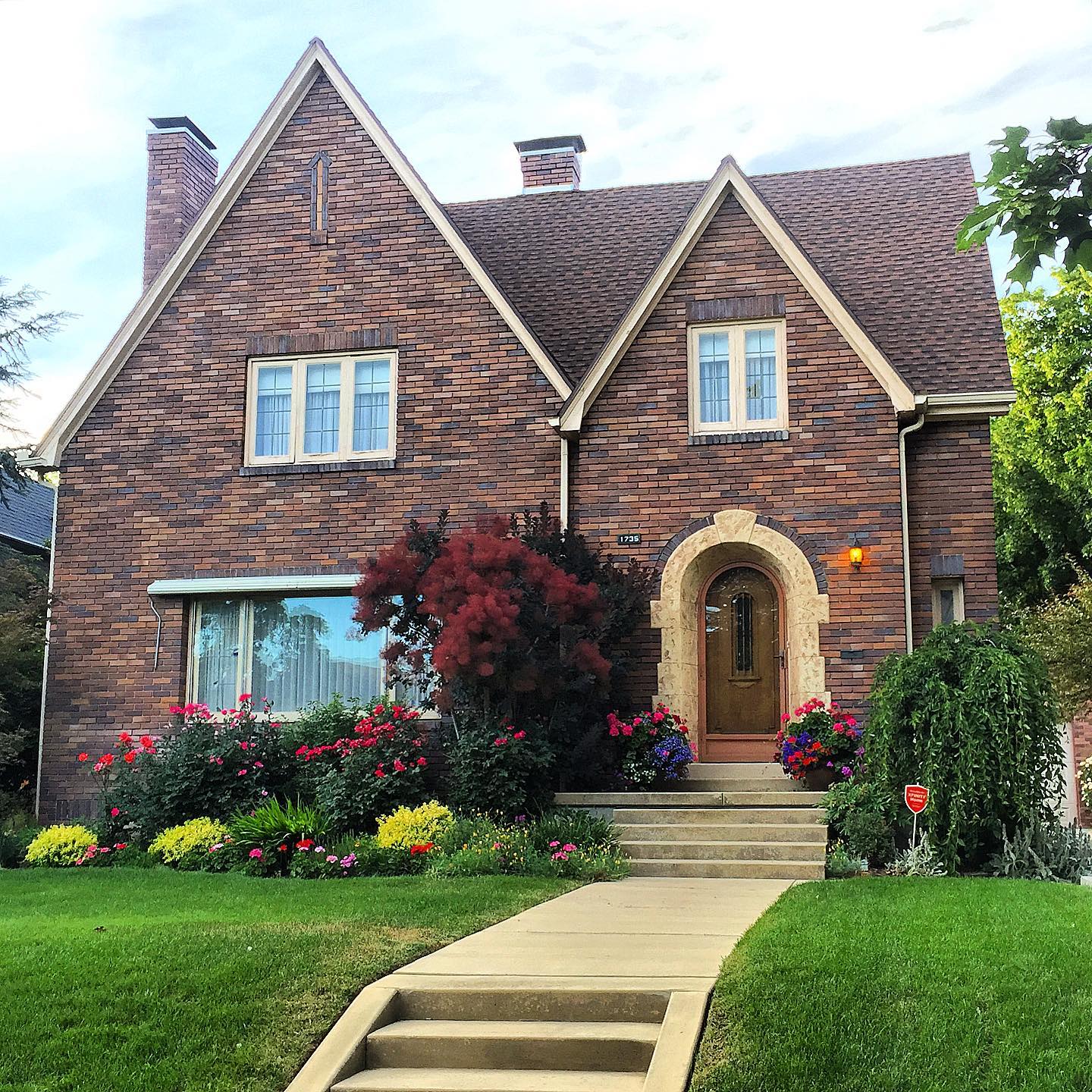 A brown brick tudor home with tan accents, a clean sidewalk and ard, and flowers blooming around the home. Photo via Instagram user @lovelyoldhomes