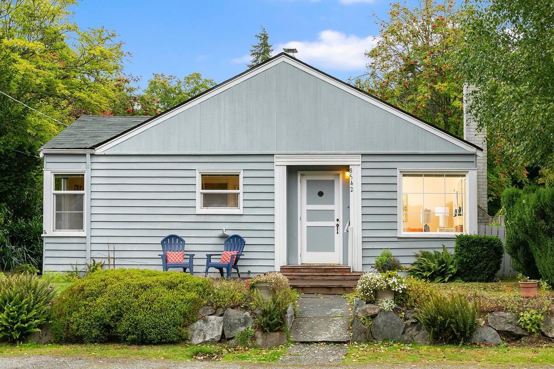 Light blue shotgun home with a front porch, two porch chairs, and a green lawn on a sunny day. Photo via Instagram user @sylviaolsby_realtor