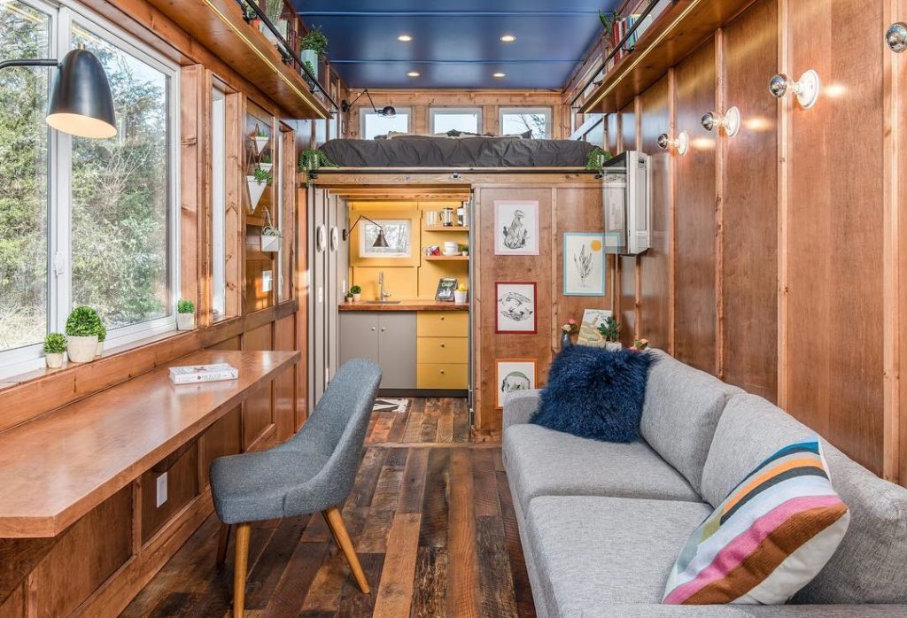 Tiny home with wood floors and wood walls and gray couches. Photo by Instagram user @newfrontiertinyhomes