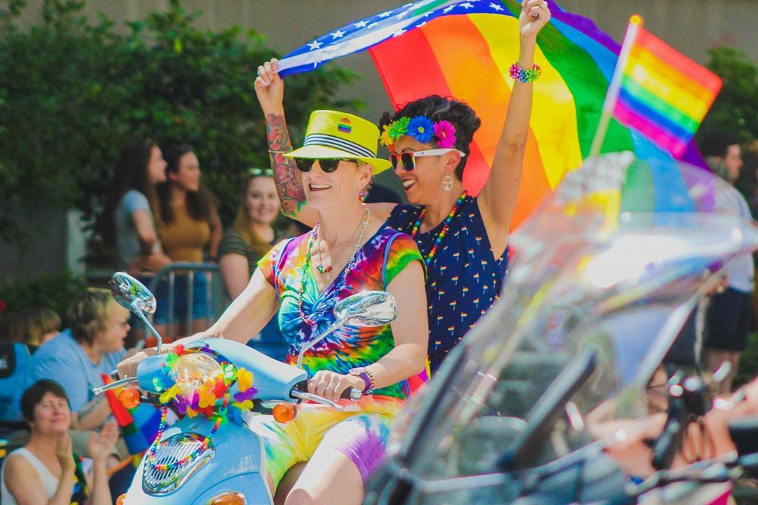 Two women holding rainbow flags riding a scooter. Photo by Instagram user @showtimechamaco