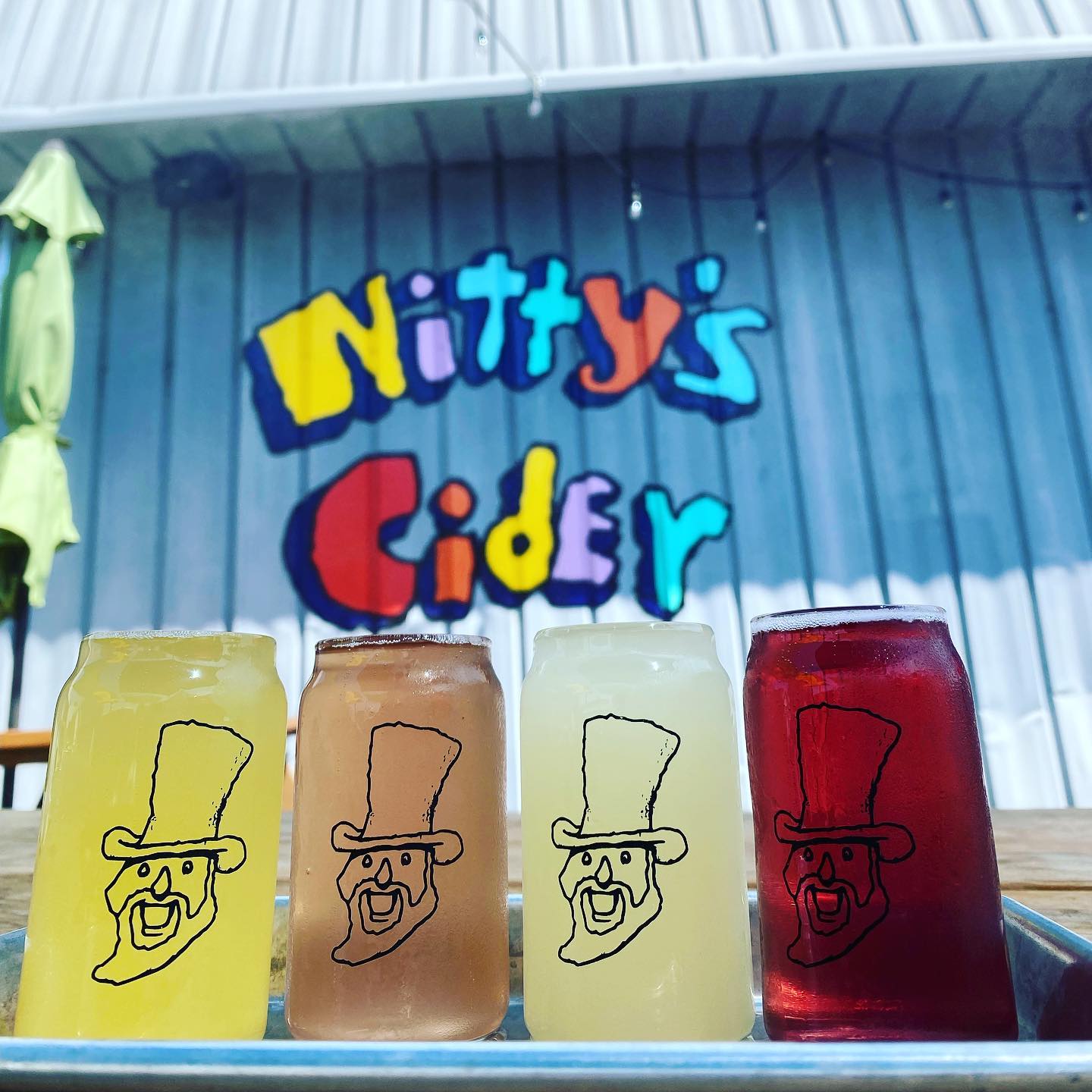 Four glasses of cider in front of the Nitty's Cider sign on a table. Photo by Instagram user @nittyscider.