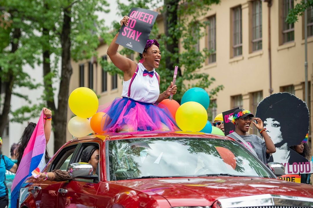A person standing though a sunroof during Sacramento Pride holding a sign that says, "Queer to Stay." Photo by Instagram user @@sacramentobee.