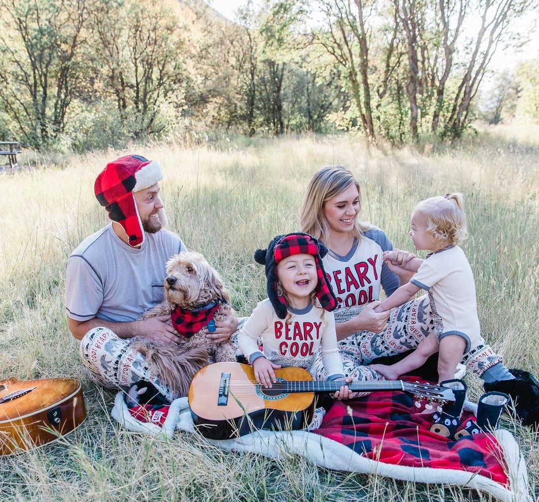 Pictured is a family portrait of a man, woman, their two children and dog sitting on grass. Photo by Instagram username @lazyoneinc
