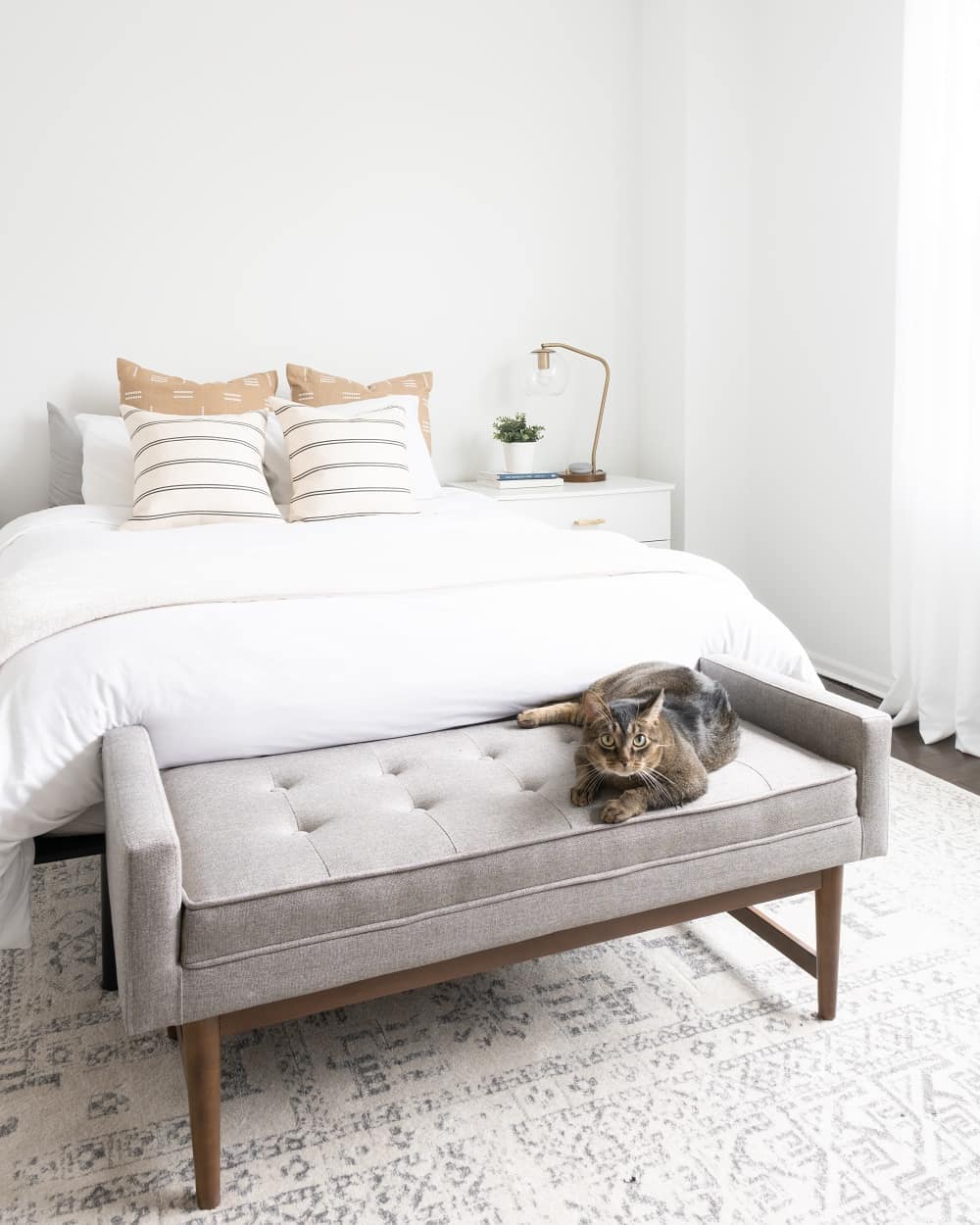 White bedroom with white bed and cat on gray bench. Photo by Instagram user @vivandtim.home