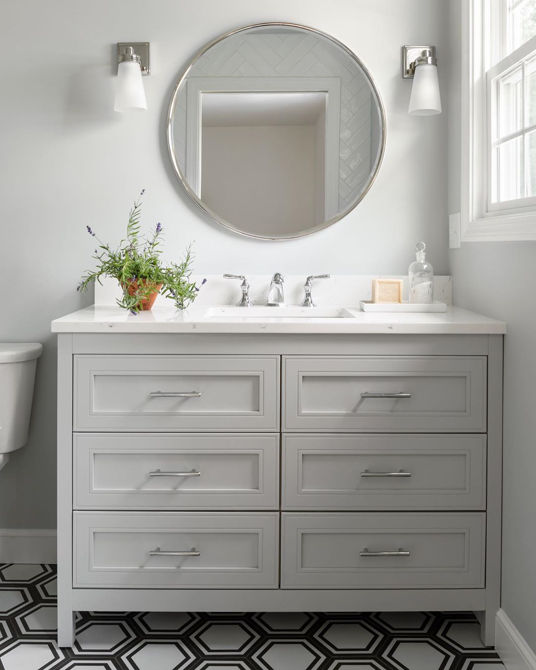 White bathroom with gray cabinets and round mirror above sink. Photo by Instagram user @tamara_flanagan_photo