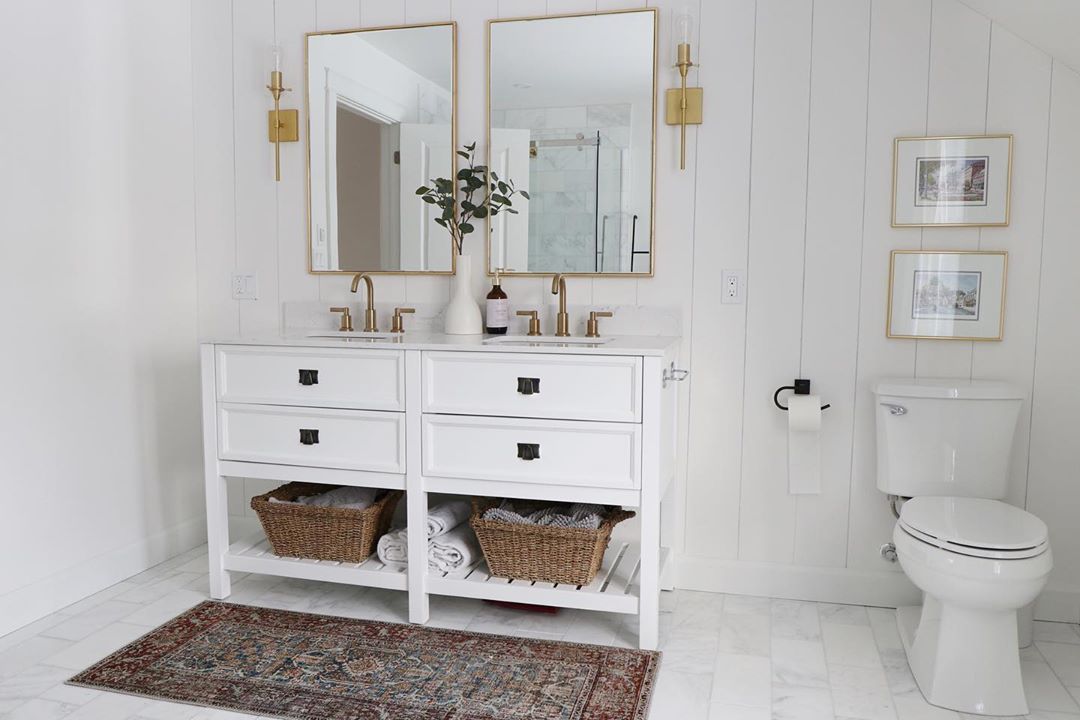 All white bathroom with gold light fixtures and wicker baskets under white cabinet. Photo by Instagram use @househappening