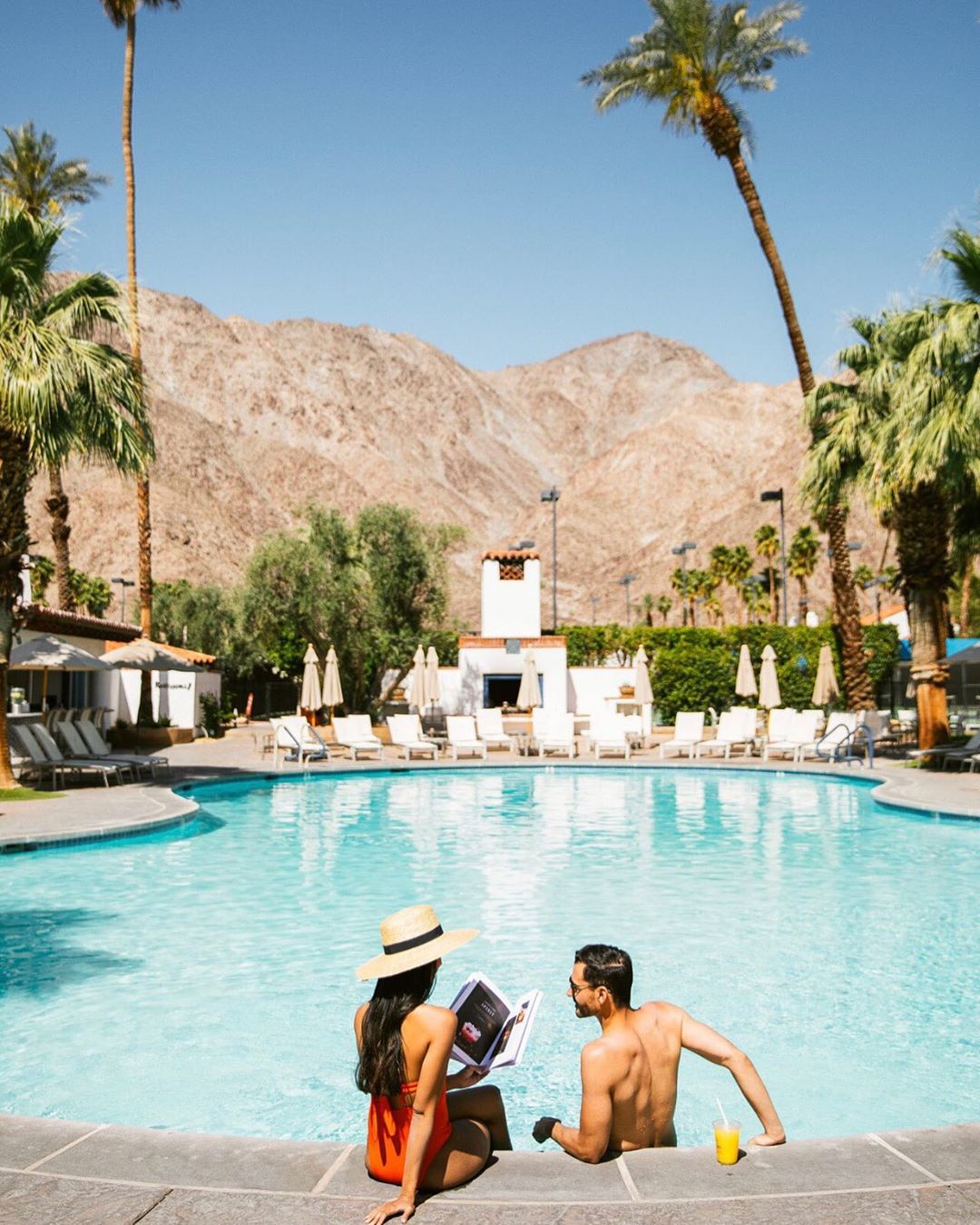 Man and woman sitting by pool with mountains in background. Photo by Instagram user @laquintaresort